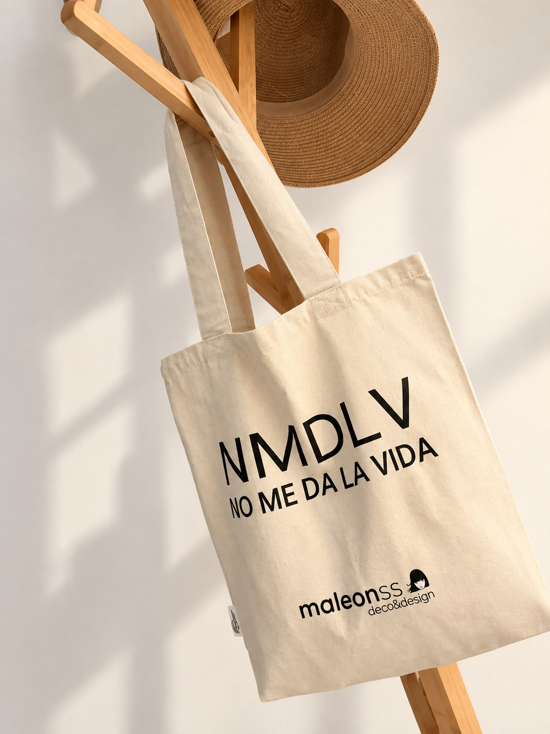 Bolsa natural con el mensaje NMDLV, No me da la vida, colgada de un perchero de madera junto a un sombrero.