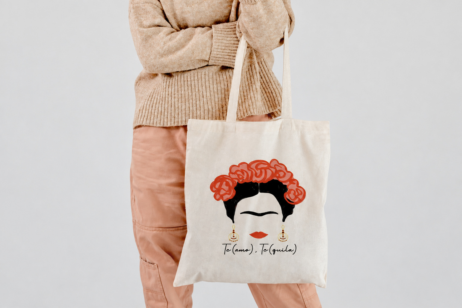Mockup de una bolsa clara de asas largas con Frida y flores rojas, llevada por una persona con jersey beige.
