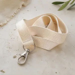 Lanyard de cinta plana marfil con mosquetón metálico sobre una superficie de piedra clara.